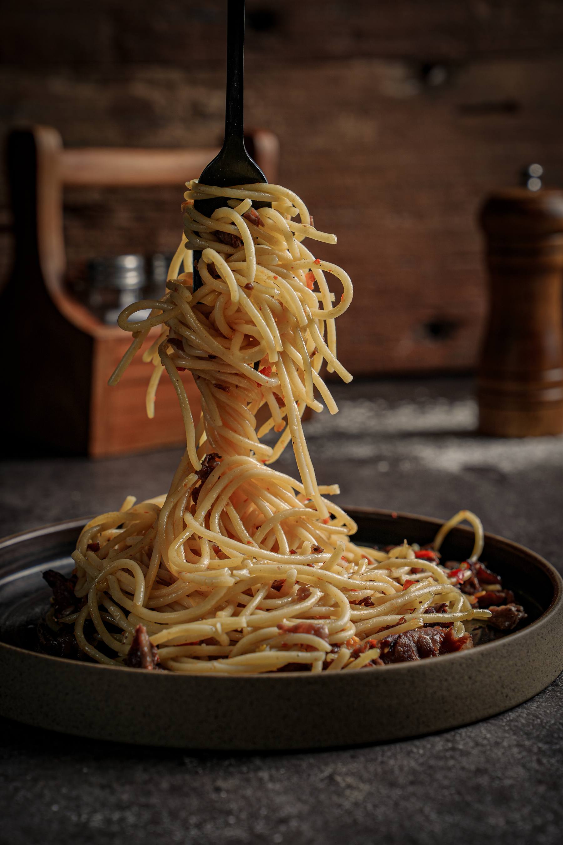 Spaghetti aglio olio lifted on a fork above a dark plate, OTSS